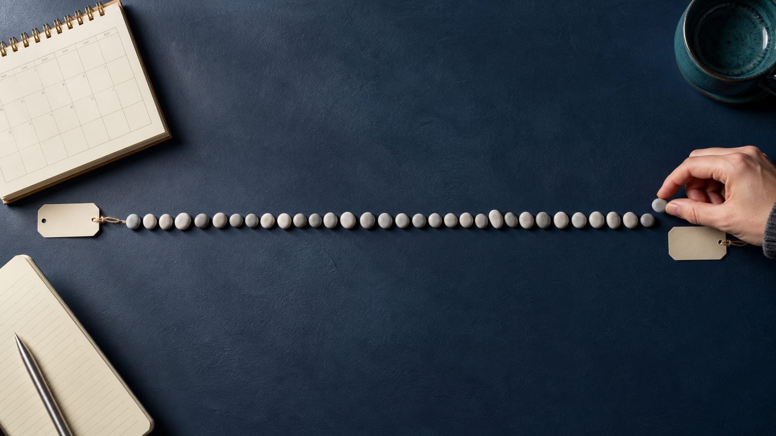 A long row of small stones forms a day-count timeline between two blank date tags, with a calendar and notebook at the edges.